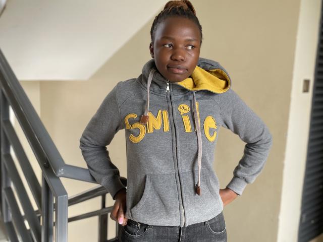 Yollah, die zelfverzekerd staat met haar handen op haar heupen, draagt een grijze hoodie met de letters "SMIC" in geel en een gele kap. met een gestreepte blouse en zwarte broek, kijkend recht in de camera. De achtergrond bevat een trap..