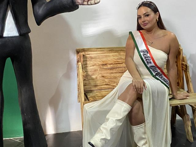 GalaMalkova, vestida con un vestido blanco sin tirantes y un cinturón rojo y verde, está sentada en una silla de madera, con las piernas cruzadas y botas blancas. de pelo oscuro, sexy, mirando la mano de un hombre que está de pie..