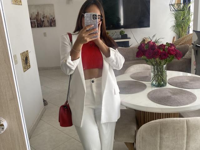 GalaMalkova se toma un selfie frente al espejo en una sala de estar moderna, vistiendo una camiseta roja corta y un blazer blanco con pantalones blancos. Sosteniendo un bolso rojo, y un jarrón con flores moradas en el fondo..
