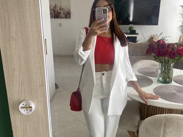 GalaMalkova, vestida con una camiseta roja y pantalones blancos, está de pie en una sala de estar moderna, sosteniendo un teléfono frente a ella. cara. Bolso rojo, televisor, jarrón con flores en el fondo..