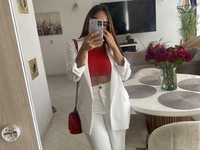 GalaMalkova, una mujer negra, está de pie en una sala de estar moderna y se está tomando un selfie frente al espejo. Vestido con un blazer blanco, un top corto rojo y pantalones blancos. Un bolso rojo en el hombro, flores rojas sobre la mesa. Se ve el pecho debajo de la chaqueta..