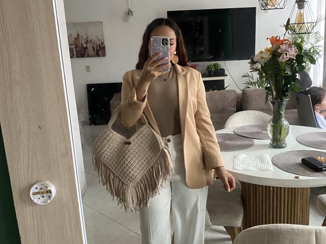 GalaMalkova è in un soggiorno moderno, tiene in mano una borsa beige con frange, indossa un blazer color sabbia e pantaloni bianchi. Si sta scattando un selfie allo specchio con il suo telefono. Composizione floreale sul tavolo.