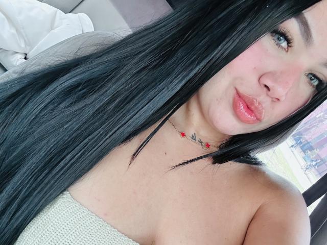 Close-up van MiaDenver, een wat dikkere vrouw met lang zwart haar, die een witte top zonder bandjes en een rode kralenketting draagt. Ze kijkt recht in de camera, met een frons en roze lippenstift..