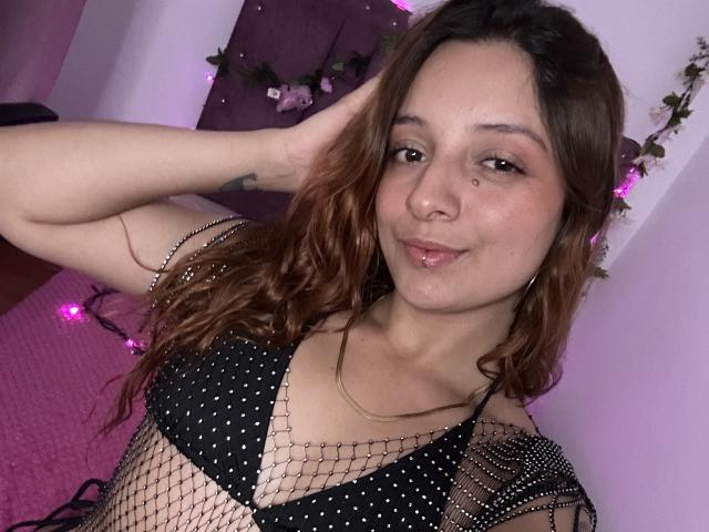 SaarahJones with long brown hair, wearing a black mesh top with polka dots, smiling slightly, hand in hair, light skin, subtle makeup, background with purple lights, casual selfie, sexy vibe.