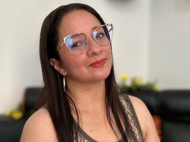 Photograph of VictoriaRousse with long black hair, wearing reflective aviator sunglasses and a gray tank top, smiling slightly with red lips. She has a confident, sexy vibe, with a subtle smirk.