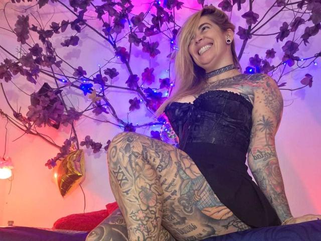 SaraValkyria, ricoperta di tatuaggi colorati, è seduta su un letto, sorridendo, indossa un corpetto nero senza spalline, con le gambe divaricate. Dietro di lei, luci al neon blu e rosa, rami di ciliegio in fiore. Molto attraente..