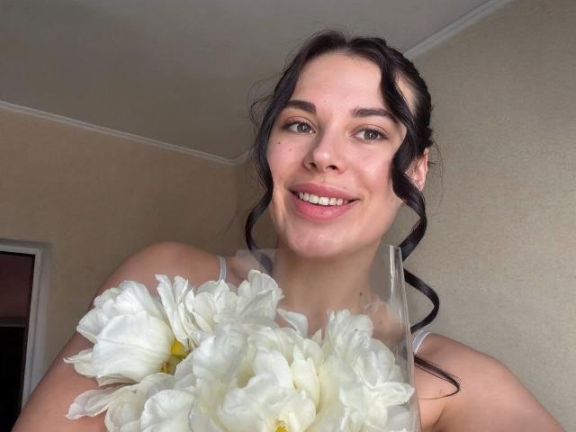 Photograph of a smiling BlackFatal with dark hair in a ponytail, holding a bouquet of white flowers, light skin, direct gaze, subtle makeup, bright indoor lighting, casual setting.