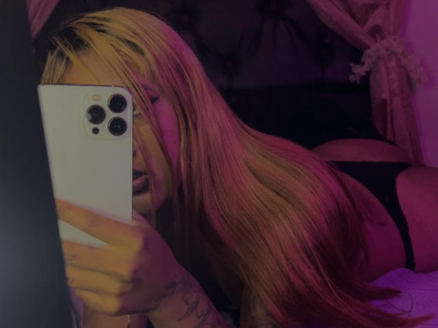NicolForest with long blonde hair takes a selfie with a white iPhone, holding it in front of her face, wearing black lingerie, lying on her stomach, pink and purple lighting.