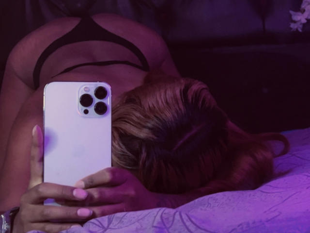 NicolForest, blonde hair, lying face down on a bed, wearing black strappy lingerie, holding a white iPhone with three camera lenses, taking a selfie, dim purple lighting, sexy, seductive pose.
