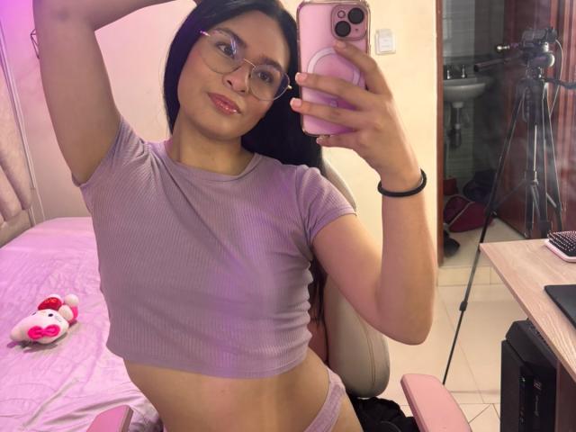 IsaDoux prend un selfie dans un miroir, vêtue d'un t-shirt violet moulant et court, ainsi que d'une culotte assortie, mettant en valeur sa silhouette mince. une silhouette mince et de petites seins. Elle tient un téléphone rose, et elle porte des lunettes..