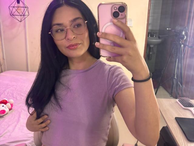 Autoportrait d'IsaDoux, une femme latina aux longs cheveux noirs, portant des lunettes et un t-shirt violet à rayures ajusté, une main sur ventre de femme enceinte, tenant un téléphone rose, chambre à coucher, éclairage rose doux..