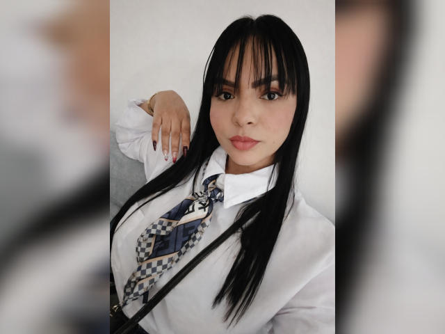 Photograph of JessicaFiore, an attractive Asian woman with long black hair, wearing a white shirt and checkered tie, looking directly at the camera with a slight smirk. Foreground blurred.