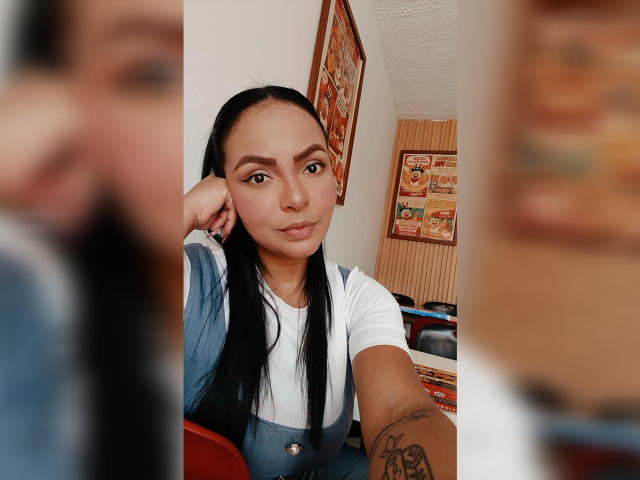 JessicaFiore, with long black hair, leans on her hand, wearing a white shirt, looking directly at the camera. Tattoo on her left forearm, blurred figure in foreground. Sexual tension, casual pose.