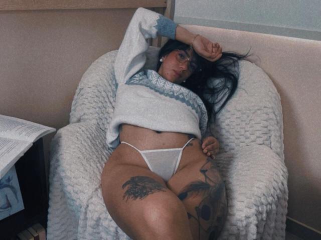 Dominatrix Melissa está recostada en un sillón blanco con textura, vistiendo un suéter gris y una tanga blanca, que deja ver tatuajes en su muslo y cadera. Su largo cabello negro se derrama sobre el reposabrazos, irradiando un atractivo relajado y sensual..