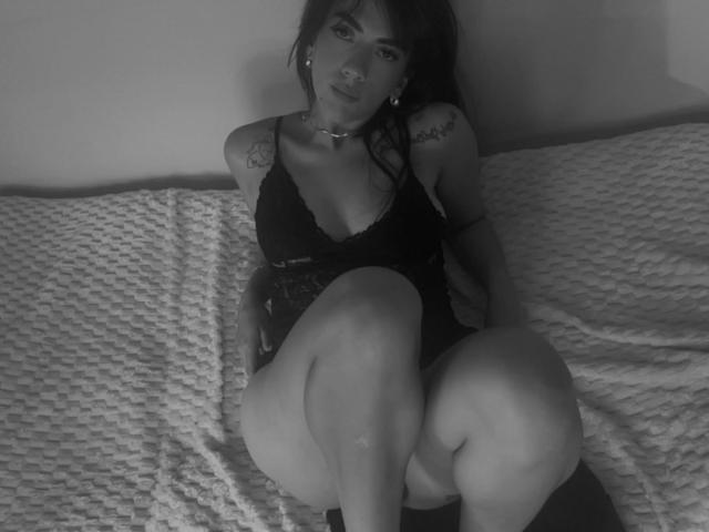 Schwarz-weißes Foto von Dominatrix Melissa, die auf einem strukturierten Bett sitzt, schwarze Spitzendessous trägt, die Beine sind gespreizt. Tätowierungen an ihren Armen und ihrer Brust sichtbar, dunkles Haar, verführerischer Blick, düstere Beleuchtung..