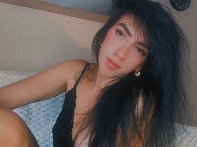 Nahaufnahme von Dominatrix Melissa, einer asiatischen Frau mit langen, schwarzen Haaren, die ein schwarzes Spitzenoberteil trägt und auf einem weißen, strukturierten gesicht, das direkt in die Kamera blickt und einen verführerischen Ausdruck zeigt. Verdammt heiß..