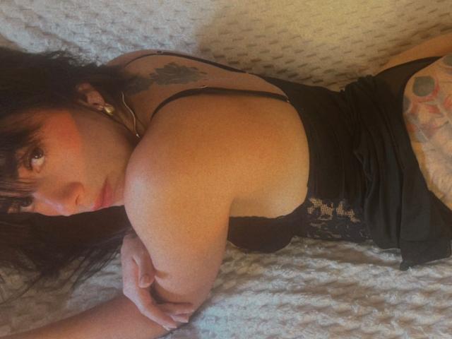 Dominatrix Melissa ligt op haar buik op een wit deken met een structuur, ze draagt een zwarte kanten korset en haar getatoeëerde arm is gekruist. Over haar borst, donker haar met een pony, een intense blik. Heel aantrekkelijk..