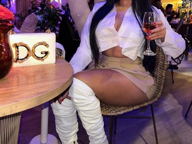 Diiana, sentada a una mesa de madera, vestía una blusa blanca anudada que dejaba ver parte de su escote, pantalones cortos beige y medias blancas hasta el muslo. botas, sosteniendo una copa de vino tinto. Escenario: un bar con poca luz..