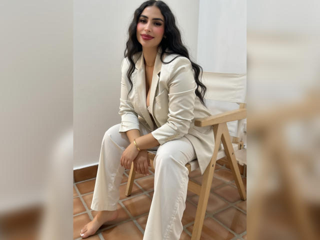 Digital artwork of MiilaWx, sitting on a wooden chair, wearing a white blazer and pants, long black hair, red lipstick, barefoot on tiled floor, bright light from window. Fucking sexy.