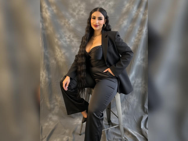 MiilaWx, a sexy woman with long black hair, sits on a stool, wearing a black strapless top and pants, black blazer, and holding a black jacket. Background is blurred grey.