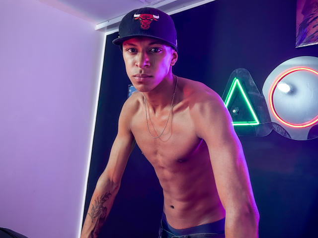 JhonatanGuzman, sem camisa, usando um boné preto com um emblema vermelho, inclina-se para frente com uma expressão séria. As luzes de neon ao fundo brilham em tons de rosa, verde e branco. Ele tem tatuagens no braço esquerdo..