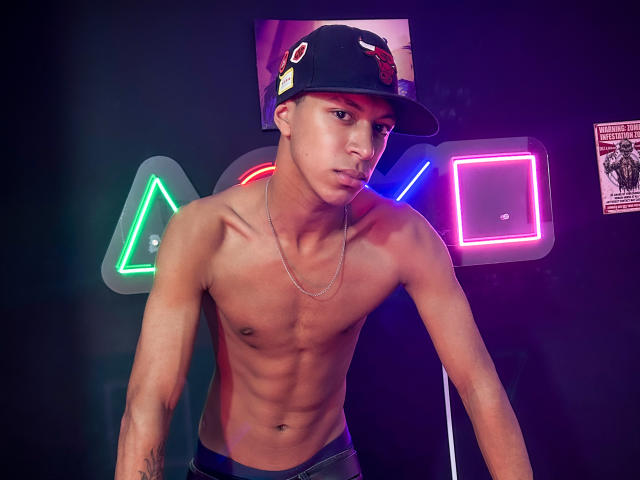 JhonatanGuzman, torse nu, musclé, portant une casquette noire avec un écusson rouge, un collier en argent, un pantalon noir, et lumières bleues en arrière-plan, regard intense, penché en avant, sexy, pose provocante..