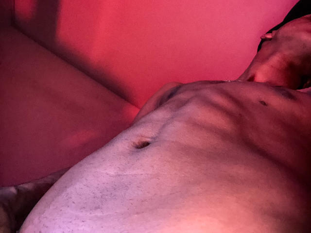 Photo en gros plan du torse nu et musclé de JhonatanGuzman, avec un abdomen bien défini, éclairé par des lumières rouges et roses. Il est allongé, la tête penchée en arrière, son pénis est partiellement visible et dissimulé par l'ombre..