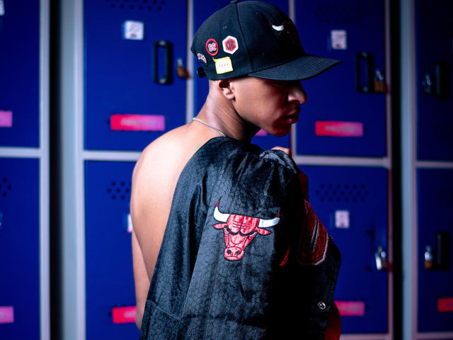Photograph of JhonatanGuzman with dark skin, wearing a black cap with red and white patches, and a sleeveless black shirt with a red bull design. Blue lockers in the background. Fucking hot.