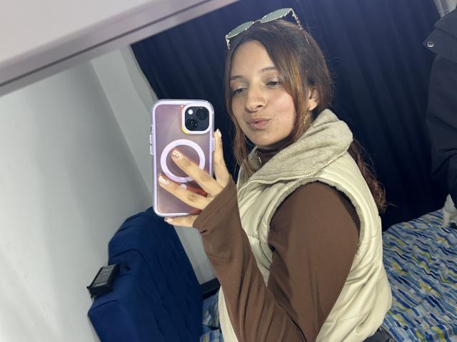 SaarahJones, holding a smartphone with a white ring grip, takes a mirror selfie. She's wearing a beige vest over a brown shirt, with sunglasses on her head. Flirty, sexy vibe.