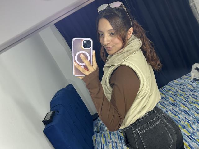 SaarahJones, volslank, draagt een bruin shirt en een gele vest, zwarte broek met hoge taille, en maakt een spiegelzelfportret met een witte telefoon. Lachend, lang bruin haar in een staart, een heup opgetrokken, seksuele spanning..