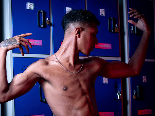 JhonatanGuzman, shirtless, muscular, dark skin, short black hair, tattoo on right forearm, chain necklace, blue lockers in background, flexing right arm, profile view, intense expression, shadowed lighting, sweaty skin.