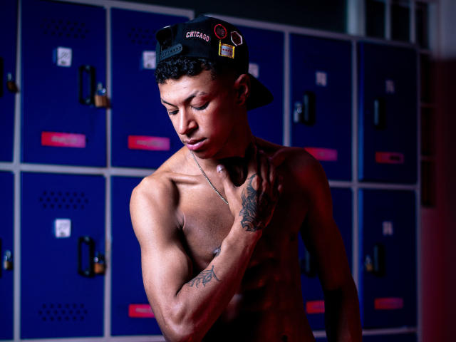 Fotografie von Jhonatan Guzman, ohne Hemd, stehend vor blauen Schließfächern. Er trägt eine schwarze Mütze, hat Tätowierungen am Arm und einen schwarzen Handschuh ohne Finger. Sein Penis ist nicht sichtbar, aber die sexuelle Spannung ist groß..
