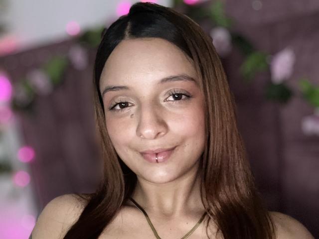 Close-up of SaarahJones, brown-haired woman with fair skin, subtle smile, visible lip piercing, brown eyes, soft lighting, pink bokeh in background, slight blush on cheeks, casual pose, sexy vibe.