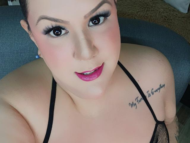 Close-up of GiiannaMorrys, fair-skinned with dark hair, heavy black eyeliner, pink lipstick, black mesh top, tattoo on right shoulder reading "Live to the Moment." Leaning forward, direct gaze, slightly flushed cheeks.