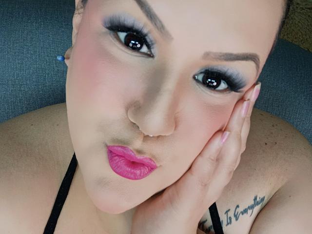 Gros plan de GiiannaMorrys, une femme à la peau claire, avec un trait d'eye-liner noir intense, du rouge à lèvres rose et des boucles d'oreilles bleues. Elle pose sa main sur sa joue, révélant un tatouage sur son épaule gauche. Extrêmement séduisant..
