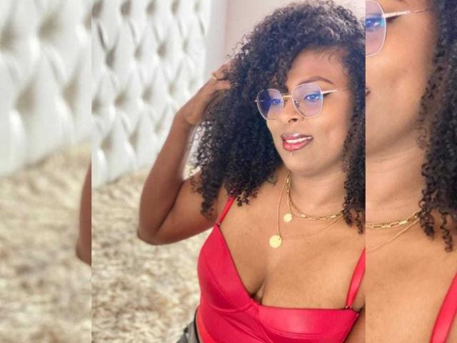 Photograph of BryannaBrown, a curvy Black woman with natural curly hair, wearing a tight red tank top, gold necklaces, and blue-tinted glasses, looking directly at the camera with a sexy expression.