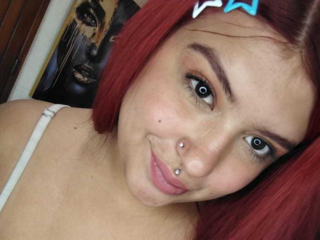 Gros plan d'AmberCutel avec des cheveux roux, un nœud bleu et un haut blanc. Elle a un piercing au nez, une petite tache au-dessus de la lèvre, et un sourire séduisant. L'arrière-plan montre un chat noir flou..