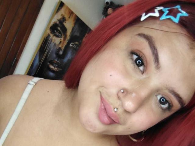 Close-up of AmberCutel with red bob, blue star hairclip, piercings on nose and lip, wearing white strap, looking at camera, cat painting in background, intense blue eyes, sexy expression.