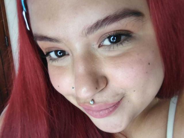 Fotografía de primer plano de AmberCutel con cabello rojo brillante, ojos azules y un piercing plateado en la nariz. Ella está sonriendo sutilmente, mostrando un rostro con pecas y un maquillaje ligero. Muy lindo y sexy..