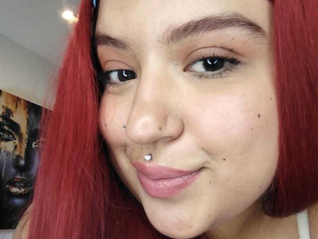 Primo piano di AmberCutel con capelli rossi, che sorride leggermente, con un piccolo piercing d'argento al naso e un piccolo piercing al labbro. anello, pelle chiara, sopracciglia scure, trucco leggero, selfie casuale.