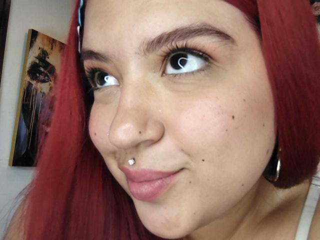 Close-up of AmberCutel with red hair, fair skin, and freckles, wearing a nose ring and hoop earrings, looking up with bright, wide eyes, slight smirk, subtle makeup, background painting.