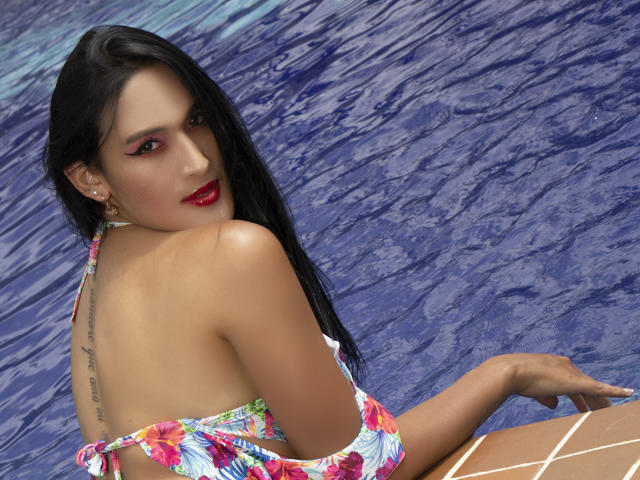 Immagine CGI di EstrellaHarryson, seduta sull'acqua blu, che guarda indietro per spalla. Ha i capelli lunghi e neri, il rossetto rosso, un costume da bagno con fiori e un tatuaggio sulla schiena. Assolutamente attraente..