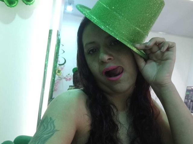 AriannaSeete wears a glittery green party hat, sticking her tongue out. She has long, dark hair, a tattoo on her left arm, and no shirt, showing her shoulders. Bright light background.
