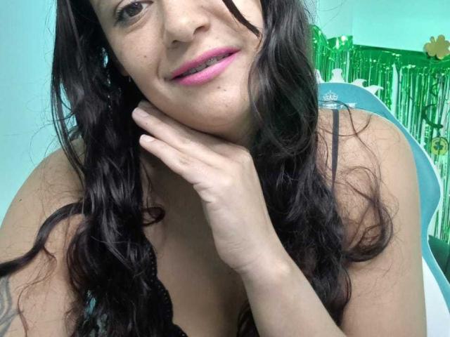 Primo piano di AriannaSeete, una donna attraente con lunghi capelli neri ondulati, rossetto rosa e pelle chiara, che appoggia la guancia su la sua mano, con del festone verde sullo sfondo. Sembra seducente..