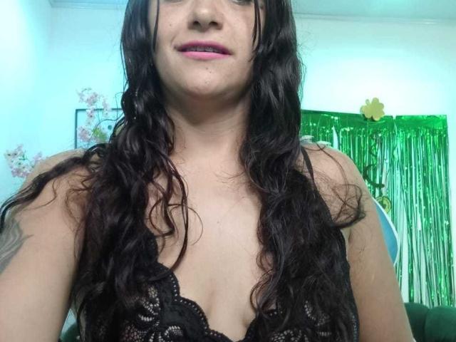 Close-up de AriannaSeete, mostrando seu rosto e parte superior do corpo. Ela tem cabelos longos e ondulados, pretos, pele clara e veste uma blusa preta de renda. Guirlanda verde ao fundo. Leve tensão sexual..