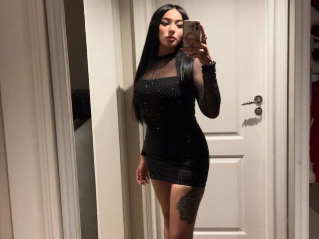 LillyTaylor, a slim woman with long black hair, stands in a doorway wearing a tight, black, sheer dress, revealing a tattoo on her right thigh. She holds a phone, taking a mirror selfie.