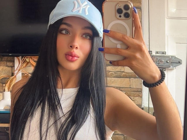 Lilly Taylor, eine attraktive lateinamerikanische Frau mit langen, schwarzen Haaren, verzog den Mund zum Kameraschwenk, während sie ein Selfie machte. Sie trägt eine weiße Mütze, ein weißes Tanktop und hat blaue Nägel. Sie hält ein iPhone..