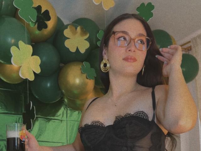 StarDustt, mit Brille und goldenen Reifenohrringen, blickt zur Seite, die Hand in der Nähe ihres Gesichts, und trägt eine schwarze Spitzenbluse. Sie steht in vorne sind grüne und goldene Ballons mit Klee-Dekorationen zu sehen. Ein Getränk halten.