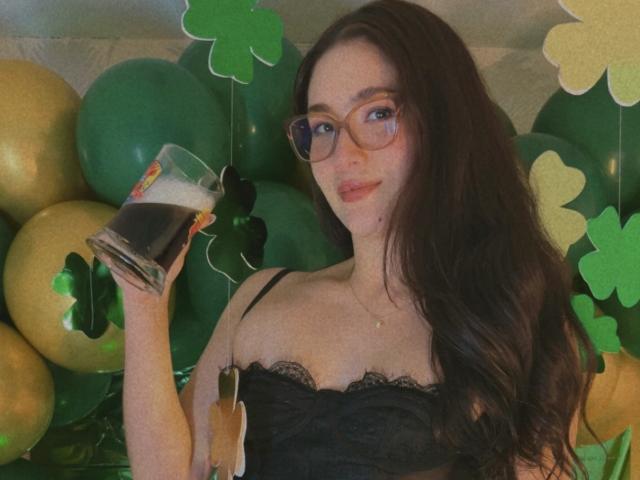 Stardustt with long black hair and glasses, wearing a black lace top, holding a pint of beer, surrounded by green and gold balloons and shamrock decorations, smirking. Fucking cute.