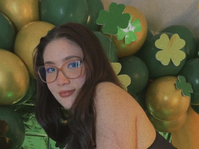 StarDustt, mit langem, dunklem Haar und Brille, lehnt sich inmitten von grünen und goldenen Ballons mit Kleeblatt-Motiven vor. Sie schaut direkt in die Kamera und hat einen leicht verführerischen Ausdruck..